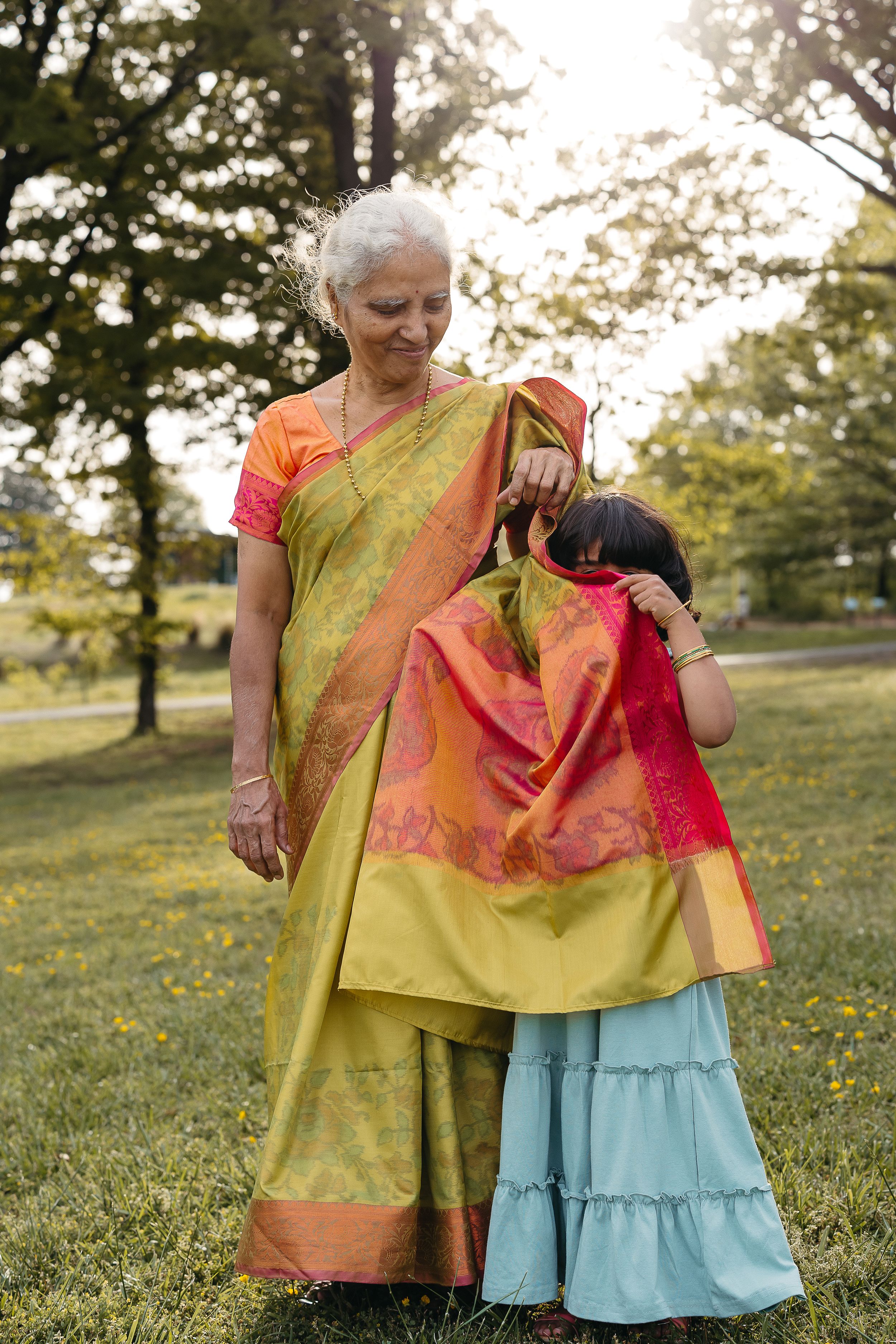 Two people in colorful traditional Indian saris stand together in a sunlit garden setting with trees in the background.