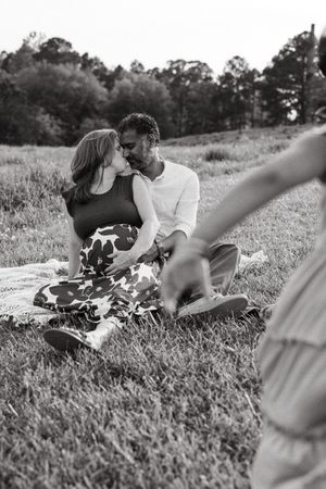 Couples share an intimate moment outdoors while sitting on a blanket in a grassy field.