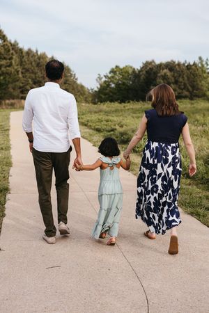 Family walking together on paved path through green meadow on sunny day with trees in background.