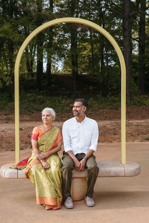 Two people sit together on a swing beneath a golden archway in an outdoor park setting.