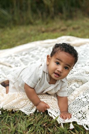 Baby in white outfit crawls on vintage lace blanket laid out on grass during outdoor photo session.