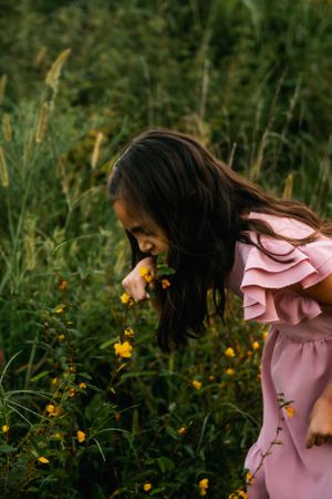 Young person in pink dress bending to smell wildflowers in a meadow.