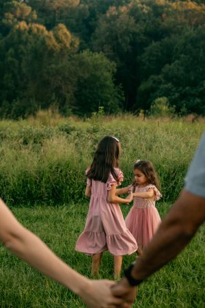 Two children in matching pink dresses play in a meadow while adults hold hands in the foreground.