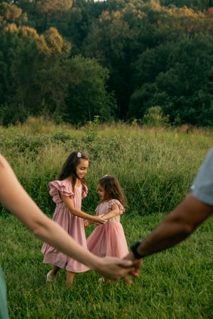 Children in pink dresses dance in a meadow while adults hold hands in the foreground.