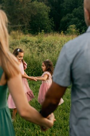 Two young children in pink dresses hold hands while playing in a grassy field as adults watch.