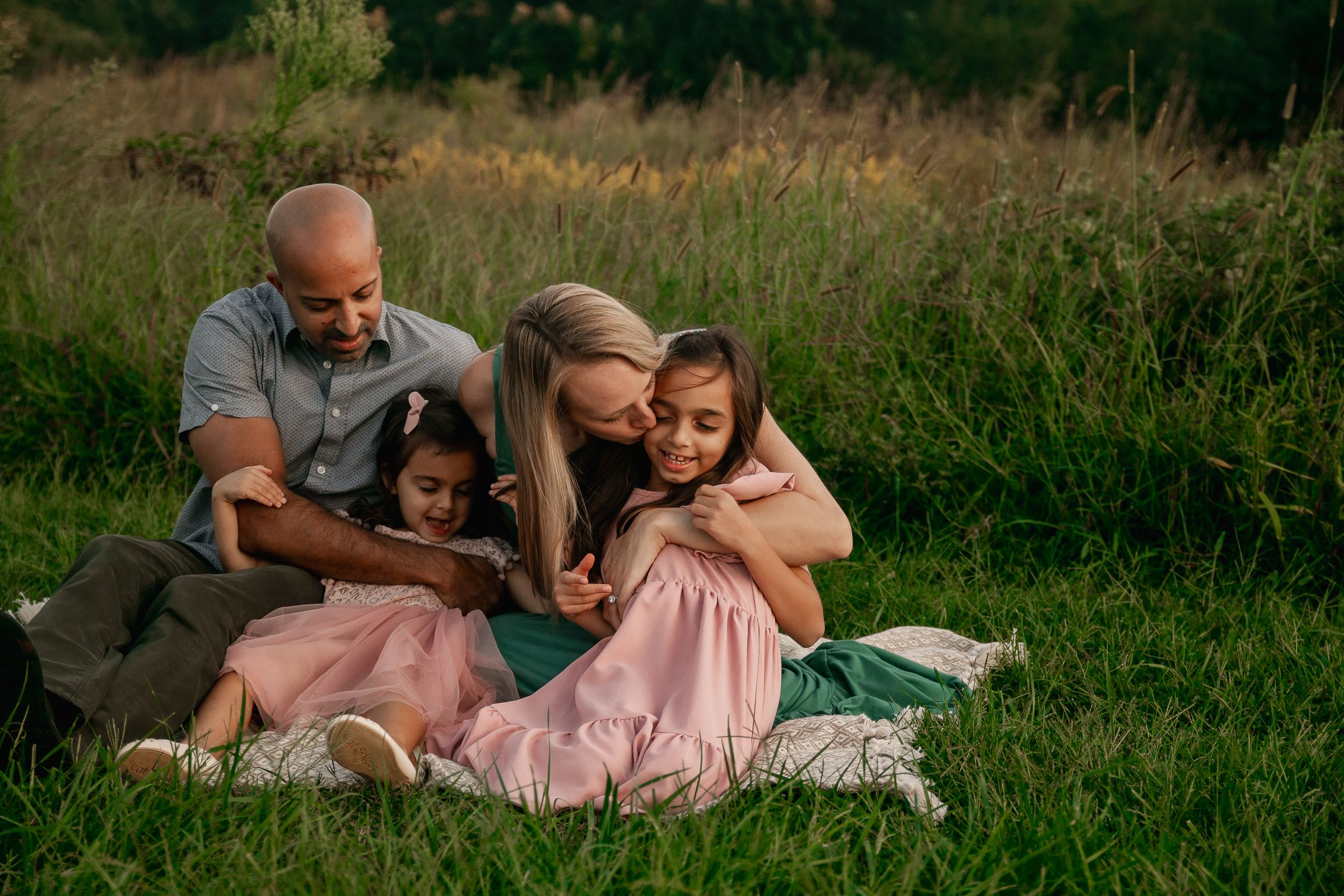 Family cuddles together on a blanket in a grassy field during golden hour.