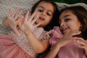 Two children in pink dresses playfully show their hands to the camera while lying down.