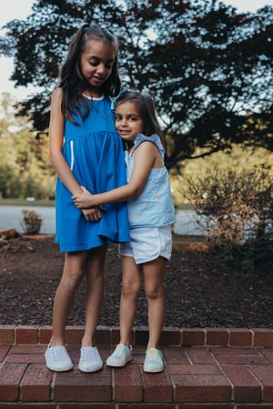 Two children in blue and white outfits stand together on a brick ledge outdoors, sharing an affectionate embrace.