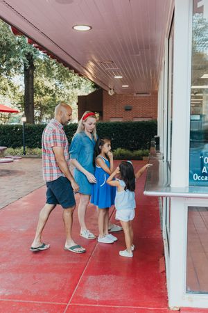 A family stands together looking at a window display on a covered red brick walkway on a sunny day.