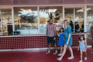 People waiting in line at a retro-style restaurant with red tiled exterior and storefront windows during sunset.