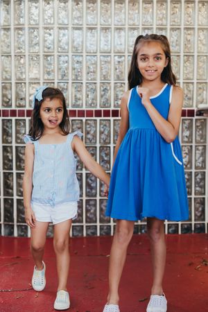 Two children in summer outfits pose together against a textured wall, wearing blue and white while standing on red ground.