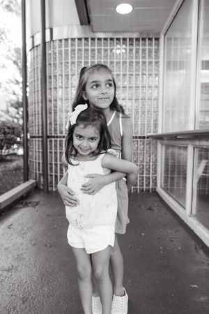 Black and white photograph of two young sisters embracing and smiling on a covered porch.
