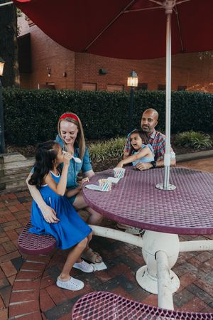 Two people sit on a brick patio at a red and white checkered picnic table under an umbrella.