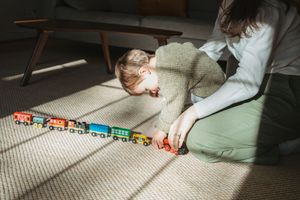 Child playing with colorful wooden toy train on carpet in sunlit room.
