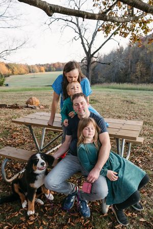 A family group sits together at a wooden picnic table with their Bernese Mountain Dog on an autumn afternoon.