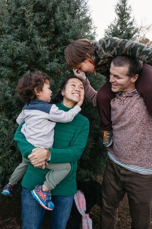 A joyful family moment outdoors with parents and two children sharing laughs against evergreen trees.
