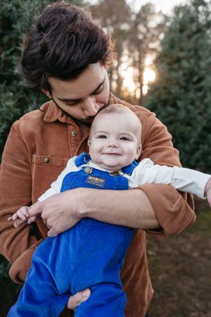 A baby wearing bright blue overalls smiles while being held in a brown jacket outdoors at sunset.