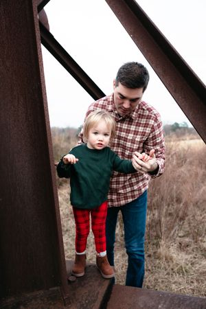 A parent and toddler stand on a rustic metal bridge, with the child wearing a green sweater and red striped pants.