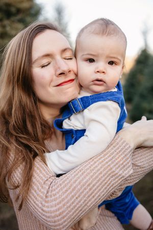 A tender moment captured outdoors as a parent in a beige sweater embraces a baby wearing blue overalls.