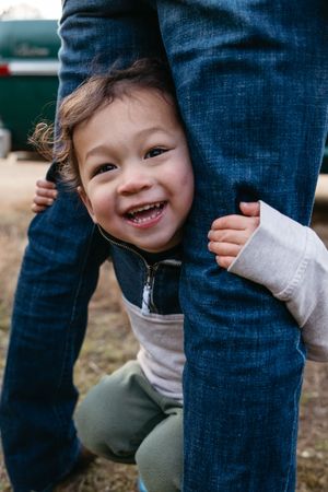 A playful child peeks out from behind legs with a bright, cheerful smile.