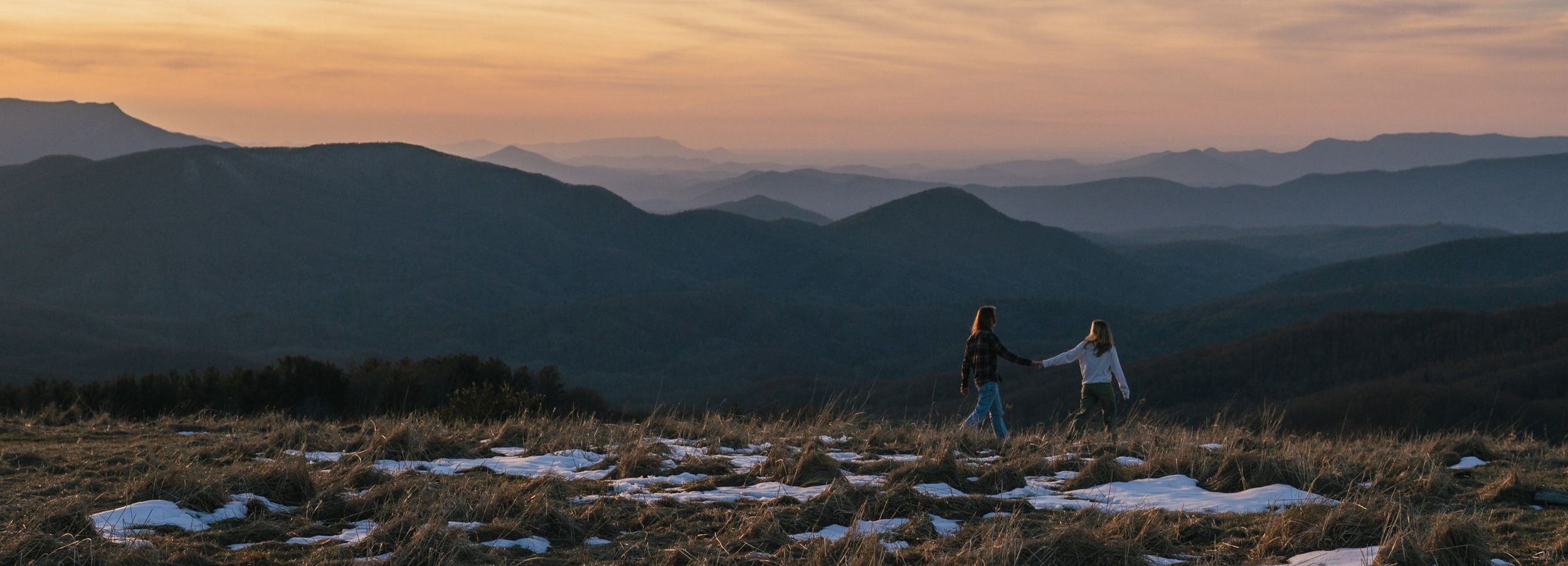 A couple walks hand in hand before a breathtaking backdrop of a sunset and mountains on Max Patch.