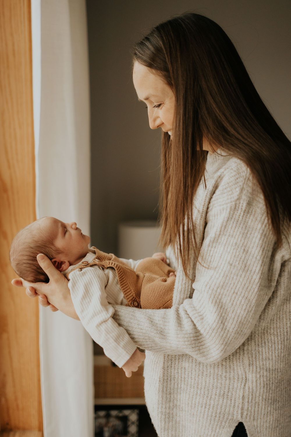 Mother in white sweater holding newborn baby by window in gentle natural light.
