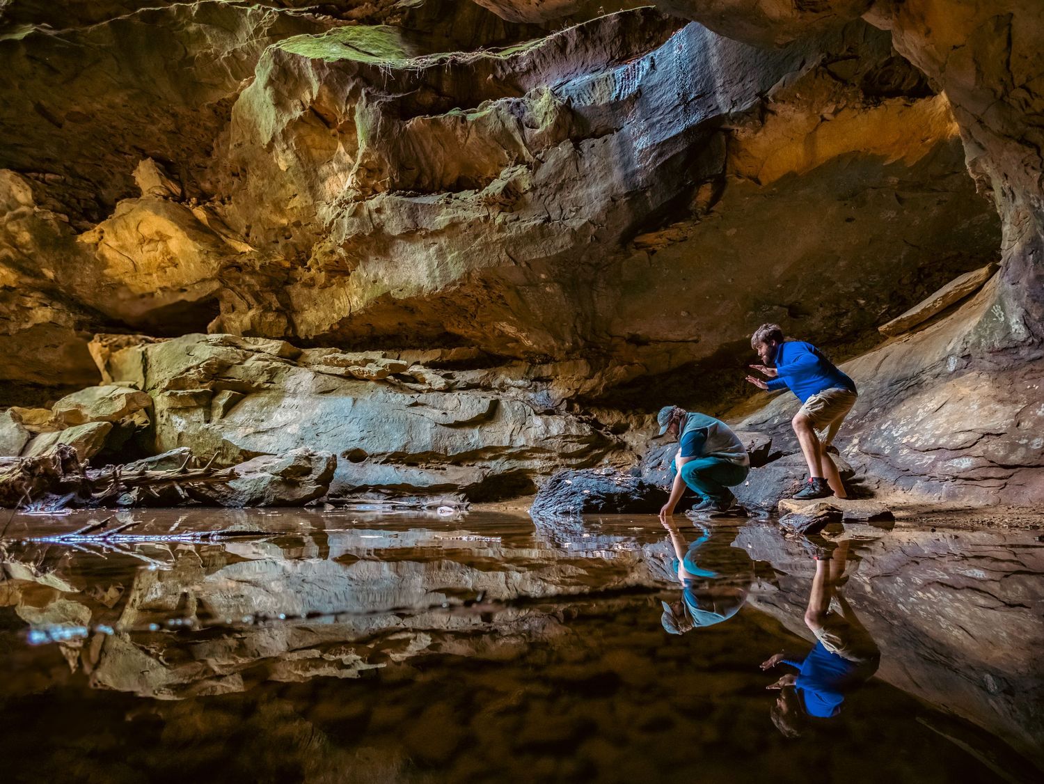 Cave explorer reflected in still underground pool with dramatic rock formations above.