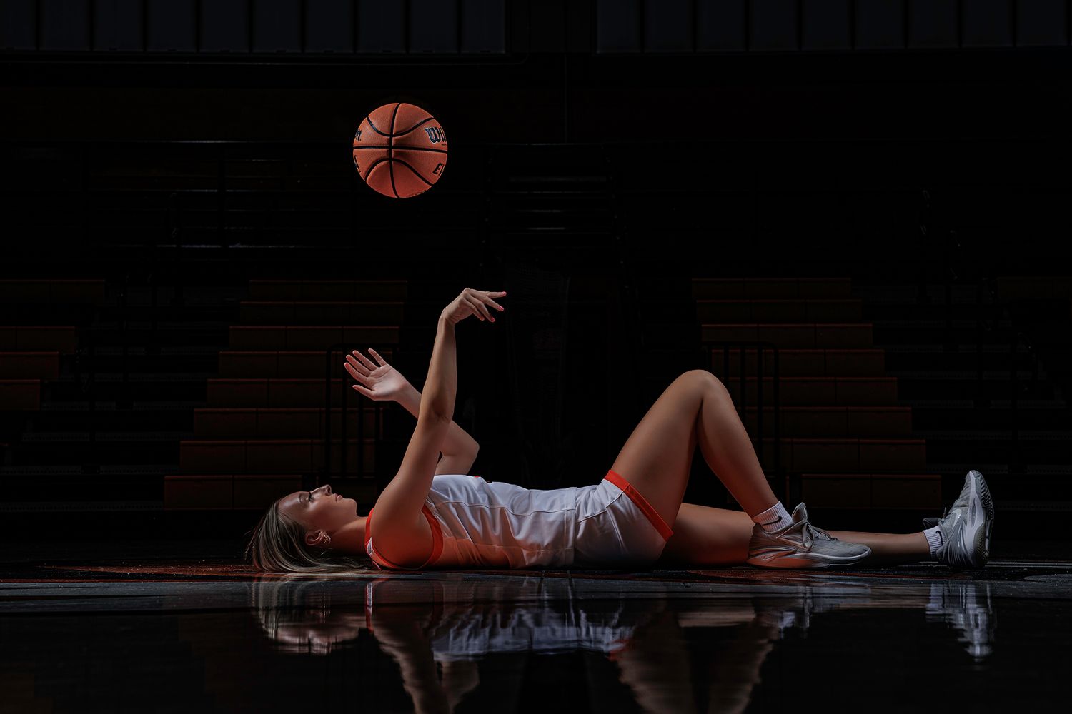 A basketball player lies on a reflective court surface tossing a ball in dramatic low-key lighting.