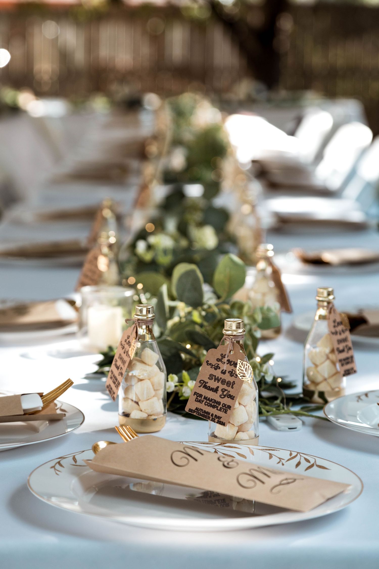 Elegant wedding table setting with white plates, gold accents, and a green eucalyptus garland centerpiece on white linens.