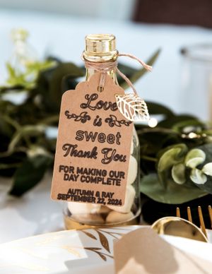 Wedding favor bottle with 'Love is Sweet Thank You' tag and decorative leaf charm against greenery background.
