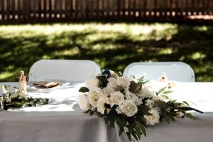 Elegant white floral centerpiece with roses decorates an outdoor table setting with white linens and chairs.