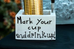 A white sign with handwritten text saying 'Mark your cup and drink up' next to a gold metallic dispenser.