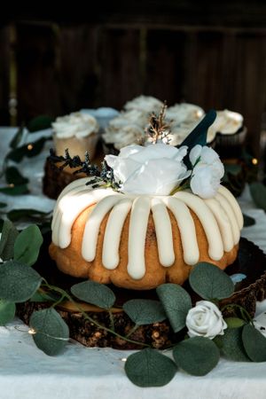 A bundt cake decorated with white icing drips, fresh flowers and eucalyptus leaves on a white tablecloth.