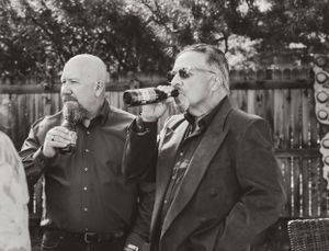 Two men in black attire drinking beverages outdoors in a black and white photograph.