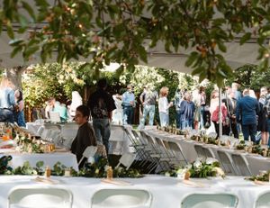 Outdoor wedding reception setup with long tables decorated with floral arrangements under tree branches at a garden venue.