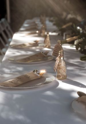 Place settings on a white tablecloth with sunlight casting shadows and glass bottles creating an elegant dining atmosphere.