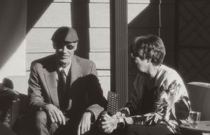 Two people sit together in a black and white photograph, one wearing sunglasses and a suit while the other is casually dressed.