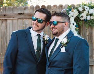 Two people in matching navy suits and sunglasses smile together at an outdoor wedding with white flowers in the background.