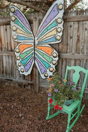 Large decorative butterfly art piece mounted on wooden fence next to a mint green rocking chair with flowers.