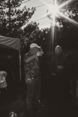 Black and white photo of two people enjoying drinks outdoors with sunlight streaming through trees.