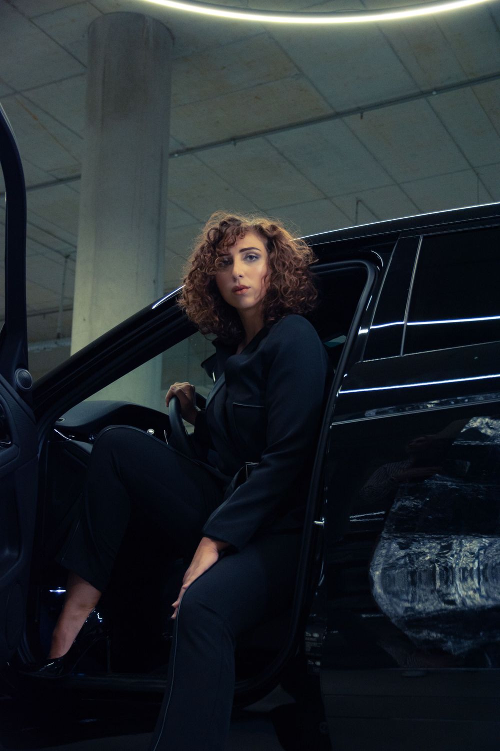 Woman in a modest black suit, sitting in a car with the door open in a Hasselt parking lot, with a cool atmosphere.