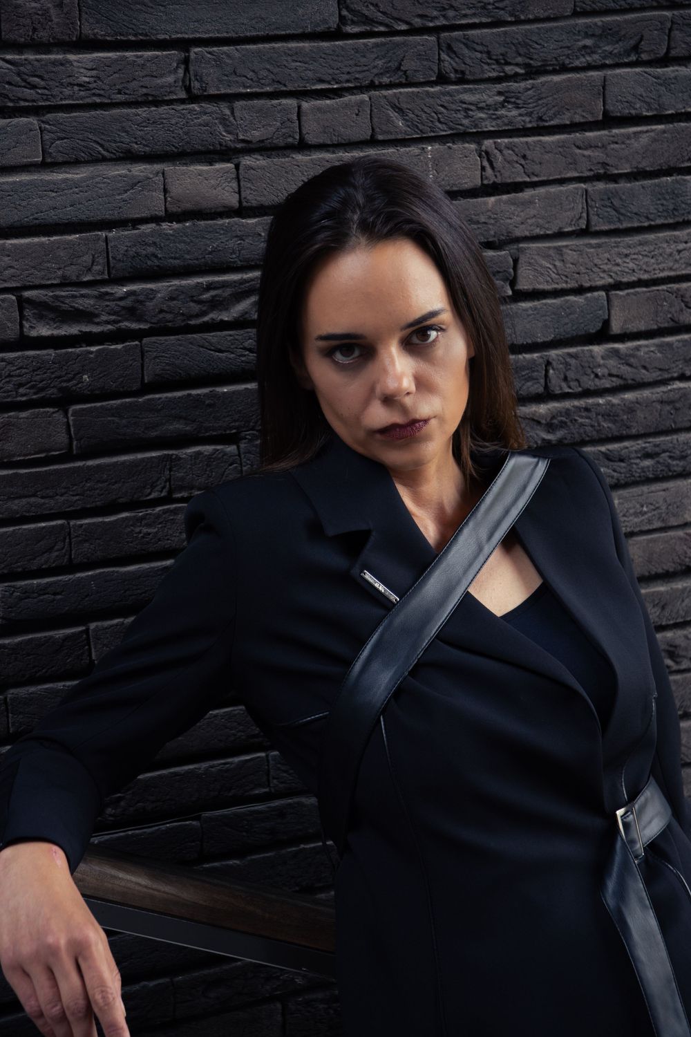 Woman in a high-fashion modest black suit, leaning against a dark brown brick wall in Hasselt, gazing fiercely at the camera.