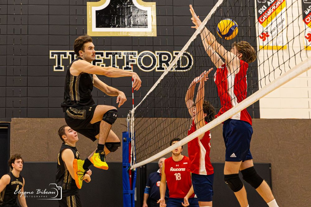 Indoor volleyball match action shows player leaping to spike ball over defenders at the net during competitive game.