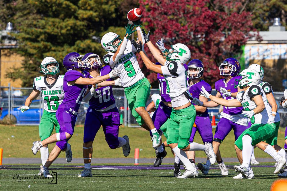 Football players in purple and green uniforms compete during a game with autumn foliage in the background.