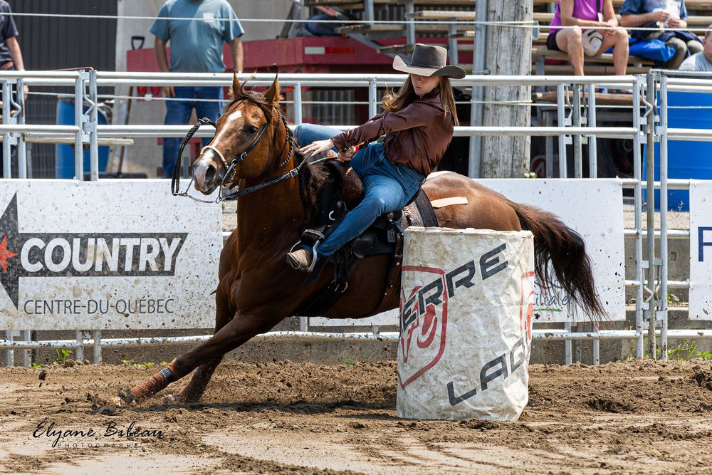 A barrel racer on horseback competes at a rodeo event, navigating around a white barrel in a dirt arena.