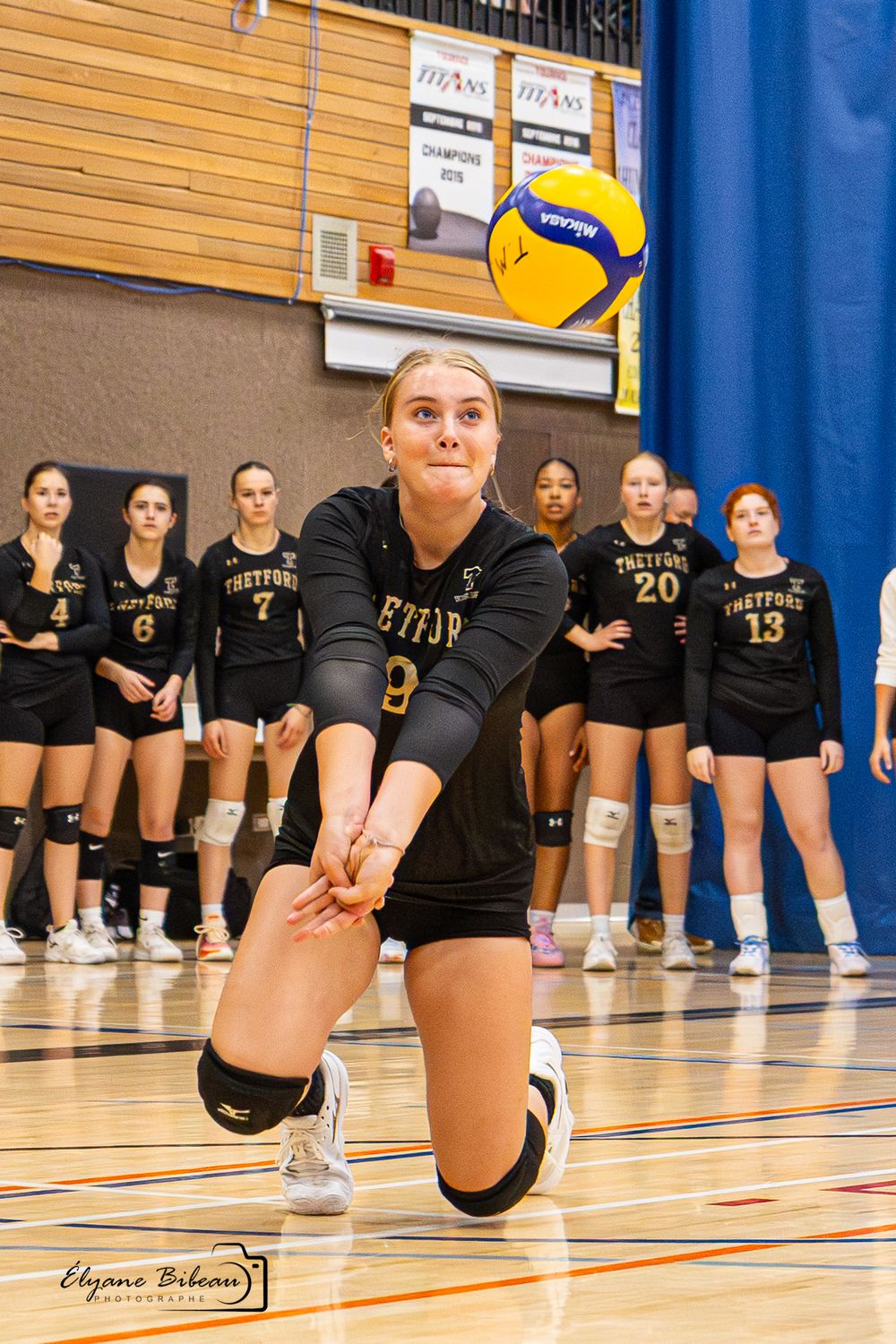 Volleyball players in black uniforms line up on a gymnasium court for a panoramic team photo.