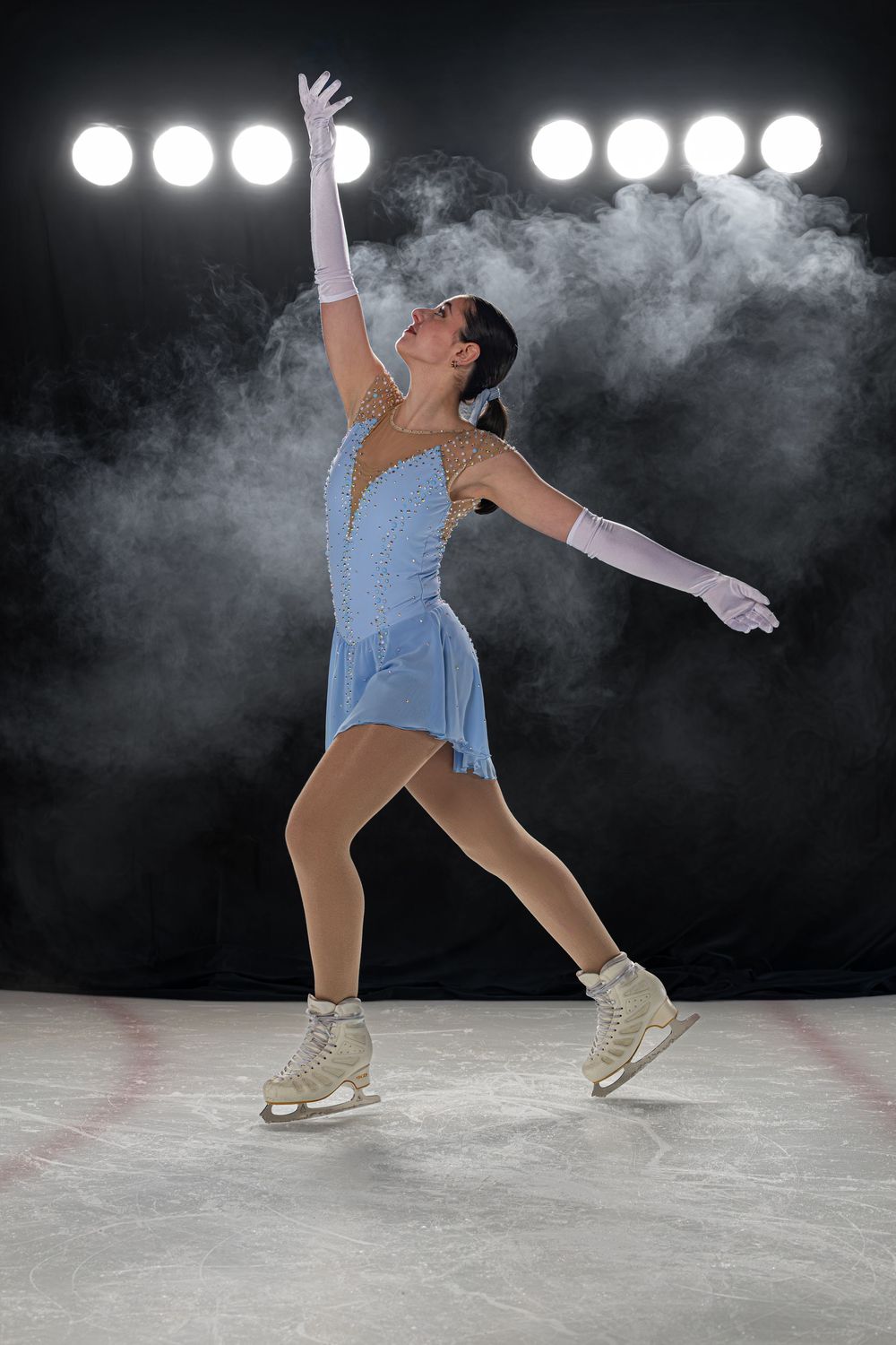 Female figure skater in blue dress and white gloves performs graceful routine on ice rink under bright stage lights.