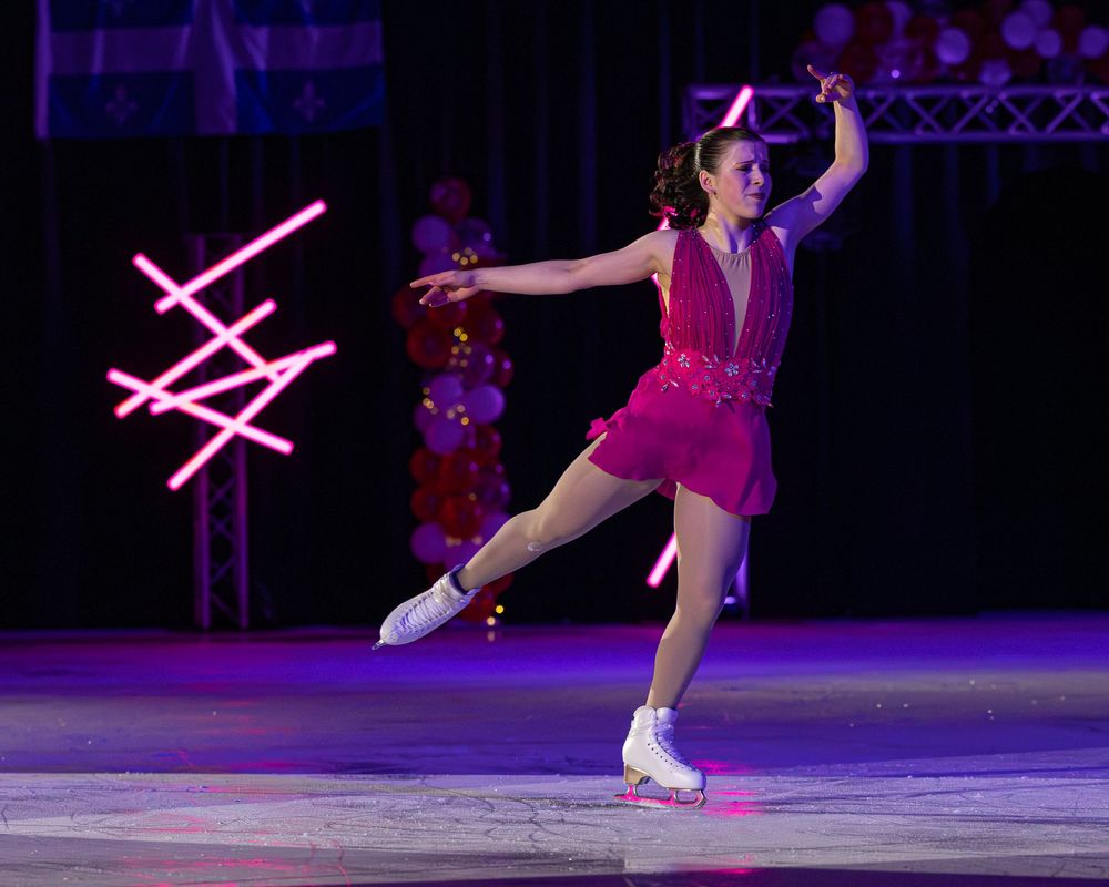 Female figure skater in pink dress performing graceful spin on ice rink with colorful neon lights in background.