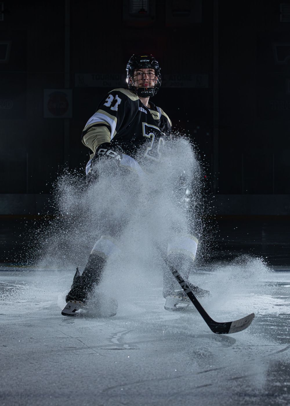 Hockey player in black jersey stops on ice creating a dramatic spray of snow under bright spotlight.
