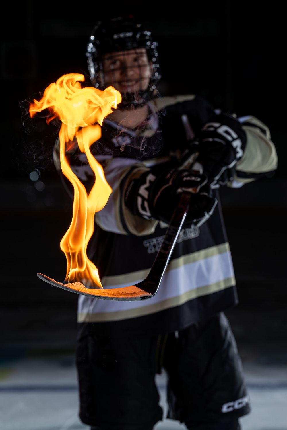 Hockey player in CCM gear holds a flaming stick blade toward camera in dramatic dark arena setting.